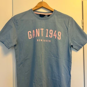 Ljusblå t-shirt från GANT med tryck - Ljusblå t-shirt från GANT med rosa tryck 'GANT 1949 NEWHAVEN' på bröstet. Klassisk rund halsringning med en rosa detalj på insidan av kragen. Perfekt för en avslappnad stil.