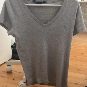 Ralph Lauren t-shirt  - ⚠️PRIS KAN DISKUTERAS⚠️Säljer en grå v-ringad t-shirt från Ralph Lauren Sport med broderad blå logga på bröstet. T-shirten är i mjuk bomull och har korta ärmar. Perfekt för en avslappnad och stilren look.