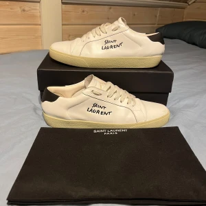 Vita sneakers från Saint Laurent - Säljer ett par vita sneakers från Saint Laurent med svart detalj på hälen och handskriven logga på sidan. Skorna har rund tå, platt sula och snörning. Perfekta för dig som gillar stilrena och exklusiva sneakers.