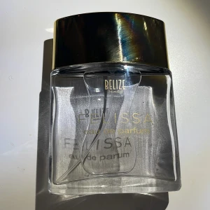 Felissa eau de parfum från Belize - En elegant parfymflaska från Belize med doften Felissa. Flaskan är genomskinlig med en stilren, rektangulär form och har ett stort, blankt guldfärgat lock. Texten på flaskan är i guld och vitt, vilket ger en lyxig känsla. Har testat den en enstaka gång.