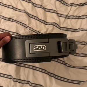 Svart träningsbälte från SBD - Säljer ett svart träningsbälte från SBD med röd insida och metallspänne. Bältet är brett och robust, perfekt för styrketräning och tyngdlyftning. Snygg SBD-logga på spännet och högkvalitativt material.