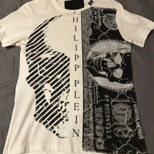 Vit t-shirt med svart mönster från Philipp Plein - Säljer en vit t-shirt från Philipp Plein med ett stort svart grafiskt mönster med stenar och text längs framsidan. T-shirten har rund hals och är tillverkad i 100% bomull. Perfekt för dig som gillar statement-plagg och unika designer.