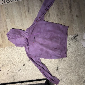 Lila hoodie från Champion - Säljer en lila hoodie från Champion med broderad logga på bröstet och loggoband längs ärmarna.