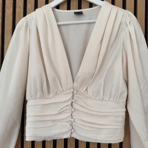 Krämvit blus med knappar från Ginatricot - Säljer en krämvit blus från Ginatricot med djup v-ringning, långa ärmar och dekorativa knappar framtill. Blusen har fina rynkade detaljer och är tillverkad i ett mjukt, lätt material.