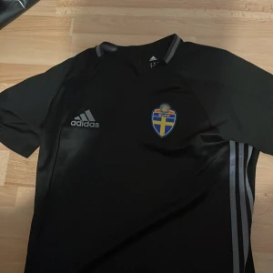 Svart Sverige fotbollströja från Adidas - Säljer en svart fotbollströja från Adidas med Sveriges landslagsmärke på bröstet och Adidas-logga. Tröjan har korta ärmar och gråa ränder på sidan. Perfekt för träning eller match!