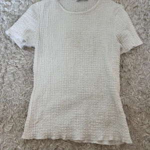 Vit strukturerad topp från Zara - Säljer en vit topp från Zara med strukturerad yta och rund halsringning. Toppen har korta ärmar och är tillverkad i ett lätt och bekvämt material. Perfekt att styla till jeans eller kjol.