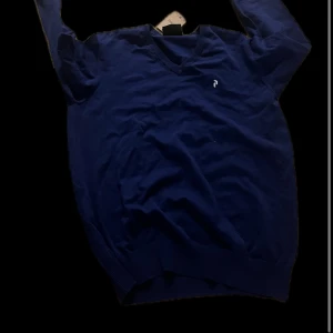 Blå stickad tröja från Peak Performance - Säljer en blå stickad tröja från Peak Performance med v-ringad hals och diskret logga på bröstet. Tröjan har långa ärmar och ribbade muddar vid ärmslut och nederkant. Perfekt för lager-på-lager under kyliga dagar.