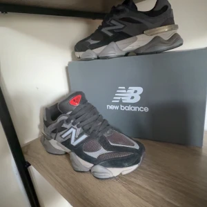 Grå New Balance sneakers - Säljer ett par grå New Balance sneakers med svarta och mörkgrå detaljer samt röd logga på plösen. Skorna har en robust sula med röda inslag undertill och klassisk snörning. Perfekta för dig som gillar streetwear och bekväma skor.