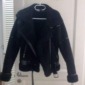 Svart bikerjacka med fuskpäls från Zara - Säljer en svart bikerjacka från Zara med fuskpäls på krage, ärmslut och nederkant. Jackan har dragkedjor, bälte i midjan. använd få tal gånger