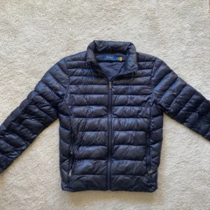 Mörkblå pufferjacka från Polo Ralph Lauren - Säljer en mörkblå pufferjacka från Polo Ralph Lauren med dragkedja och ståkrage. Jackan har broderad logga på bröstet och två fickor med dragkedja. 