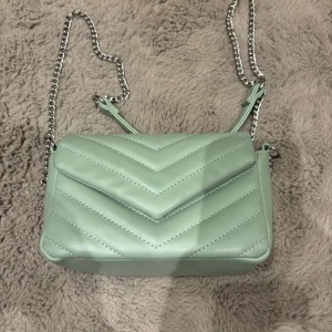 Ljusgrön quiltad axelväska med kedja - Säljer en ljusgrön axelväska med quiltat mönster och silverfärgad kedjerem. Väskan har ett stilrent V-mönster på locket och flera fack inuti. Perfekt för dig som vill ha en snygg och trendig accessoar till din outfit. Ps lilla fläcken är borta!