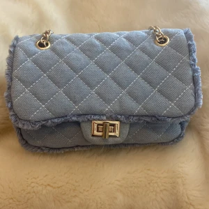 Ljusblå quiltad axelväska med kedja - Säljer en ljusblå quiltad axelväska med guldfärgad kedja och vridlås. Väskan har mjukt tyg med fransade kanter och rutmönster. Perfekt för att lyfta din outfit med en trendig touch.