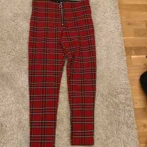 Röda rutiga byxor med dragkedja från Zara Basic - Snygga röda byxor från Zara Basic med klassiskt rutigt mönster och en stor silverfärgad dragkedja framtill. Byxorna har hög midja och smal passform, perfekta för att sticka ut i mängden.
