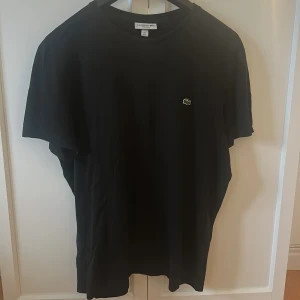 Svart t-shirt från Lacoste - Säljer en klassisk svart t-shirt från Lacoste med den ikoniska krokodilloggan broderad på bröstet. T-shirten har rund halsringning och normal passform. Perfekt basplagg för garderoben!