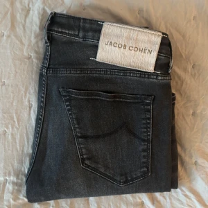 Svarta jeans från Jacob Cohën - Stilrena och tidlösa jeans i mycket bra skick. Modellen har en smal passform och är tillverkad i högkvalitativt material med exklusiv patch med logotyp baktill i midjan.  Storlek 34/W30,Passformen är slimmad utan att vara för tight – jag är 187 cm lång och väger 68 kg, och byxorna sitter snyggt och bekvämt på mig. Perfekta jeans för dig som uppskattar kvalitet och en klassisk stil. Nypris 5500 kr, nu endast 1799 kr   Har du några frågor eller vill ha fler bilder? Skicka gärna ett meddelande! 