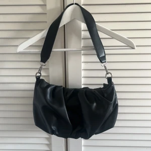 Svart axelväska i syntet - Trendig svart axelväska med bred axelrem och rynkad design. Väskan har silvriga metalldetaljer och är tillverkad i ett slätt syntetmaterial. Perfekt för att lyfta din outfit med en modern touch.
