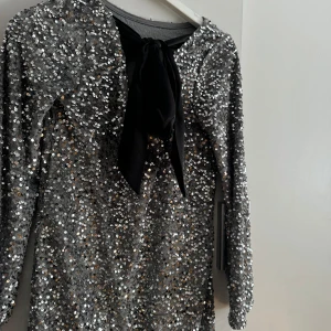Glittrig silverklänning med rosett - Säljer en glittrig silverfärgad klänning med långa ärmar och svart rosett i bak. Klänningen är täckt av paljetter, endast använd en kväll och inga defekter!