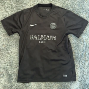 Svart PSG x Balmain fotbollströja från Nike - Säljer en svart PSG x Balmain fotbollströja från Nike med vitt tryck på bröstet och Dri-Fit-material. Tröjan har korta ärmar, rund hals och autenticitetsmärke från 2021. Perfekt för dig som gillar stilrena matchtröjor.