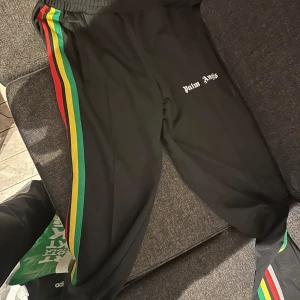 Svarta träningsbyxor med färgglada ränder från Palm Angels - Svarta träningsbyxor från Palm Angels med gröna, gula och röda ränder längs sidorna. Byxorna har resår i midjan och logotypen broderad på ena benet. Perfekta för en sportig och trendig look.