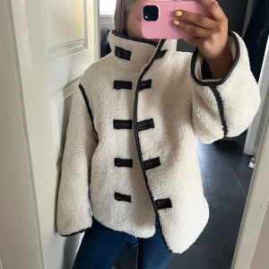 Vit teddyjacka med svarta detaljer - Säljer en vit teddyjacka med svarta kantdetaljer och dekorativa knappar framtill. Jackan har hög krage och lång ärm, perfekt för kyligare dagar. Snygg och trendig modell som sticker ut med sina kontrasterande detaljer.