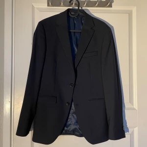 Mörkblå kostym med kavaj och byxor - Säljer en stilren mörkblå kostym med matchande kavaj och byxor. Kavajen har två knappar framtill, klassisk krage och fickor med lock. Byxorna har pressveck och bakficka med knapp. Perfekt för formella tillfällen. Använd två gånger 