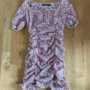 Blommig kortklänning med volanger - Supersöt kortklänning med rosa och grönt blommönster på vit botten. Klänningen har korta puffärmar, volangdetaljer och rynk längs hela framsidan. Perfekt för sommarens alla tillfällen!