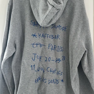 Grå hoodie från Samsøe Samsøe - Grå hoodie från Samsøe Samsøe med blå text och grafiskt tryck på ryggen samt liten logga framtill. Klassisk huva och känguruficka. Perfekt för en avslappnad stil. Passar även L.