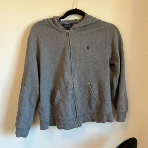 Grå hoodie från Polo Ralph Lauren - Säljer en stilren grå hoodie från Polo Ralph Lauren med dragkedja och broderad logga på bröstet. Perfekt för en avslappnad look. Den har långa ärmar och en praktisk huva. Den är i fint skick, skriv om minsta lilla fundering!