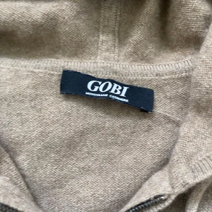 Beige hoodie i kashmir från GOBI - Mysig beige hoodie i mjuk mongolisk kashmir från GOBI. Tröjan har dragkedja framtill, huva med dragsko och två fickor. Perfekt för en avslappnad och stilren look.