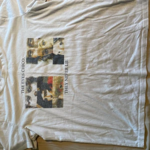 Vit t-shirt med tryck och text - Vit t-shirt med grafiskt tryck av ansikten och texten 'THE EYES CHICO, THEY NEVER LIE' på framsidan. Klassisk passform och rund hals. Perfekt för dig som gillar unika prints och stilrena plagg.