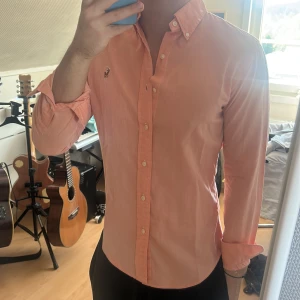 rosa skjorta från Ralph Lauren - Säljer en laxrosa skjorta från Ralph Lauren med klassisk button-down krage och broderad logga på bröstet. Skjortan har långa ärmar och vita knappar framtill. Perfekt för en stilren look.