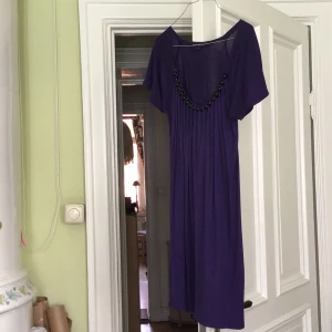 Lila klänning med pärldetaljer från French Connection - Säljer en lila klänning från French Connection med korta ärmar och dekorativa svarta pärlor längs ringningen. Klänningen har en rak passform och är knälång, perfekt för dig som gillar färg och unika detaljer.