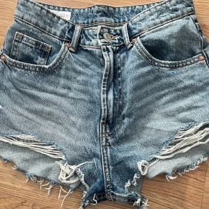 Blå jeansshorts med fransar från ZARA - Säljer ett par blå jeansshorts från ZARA i storlek XS. Shortsen har råa fransade kanter och slitna detaljer framtill, klassisk femficksmodell och knappgylf. Perfekta för varma dagar.