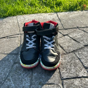 Svarta sneakers med färgglad sula - Säljer ett par svarta sneakers med vit snörning och kardborreband. Insidan är röd och sulan har ett färgglatt, graffitiliknande mönster. Skorna har rund tå och platt sula, perfekta för dig som vill sticka ut lite extra.