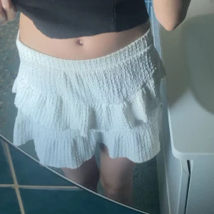 Vit volangkjol med struktur - Säljer en söt vit kjol med volanger och strukturerat tyg. Kjolen är kort och har en luftig känsla. Har en liten liten fläck som knappt syns 