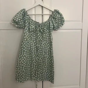 Grön blommig kortklänning med puffärmar - Superfin ljusgrön klänning med vita blommor och puffiga kortärmar. Klänningen har knytning framtill vid bysten och en söt, luftig passform. Perfekt för vår och sommar!