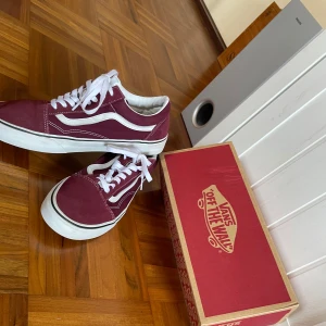 Vinröda Vans Old Skool sneakers - Säljer ett par vinröda Vans Old Skool sneakers med vit sula och klassiska vita sidoränder. Skorna har snörning och är i en snygg färg som sticker ut. Perfekta för dig som gillar streetstyle och vill ha något bekvämt och stilrent.