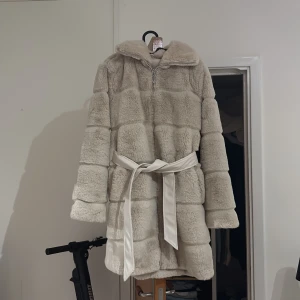 Beige pälsjacka från Amisu med bälte - Säljer en beige pälsjacka från Amisu i storlek 34. Jackan har lång ärm, dragkedja framtill och ett matchande bälte i midjan. Perfekt för kyliga dagar när du vill vara både varm och snygg. Bra skick använd 2 gången 