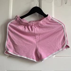 Rosa sportiga shorts med vit kant - Säljer ett par ljusrosa sportiga shorts med elastisk midja och vita kanter längs sidorna och bensluten. Perfekta för träning eller varma dagar. Mjukt bomullsmaterial och klassisk passform.