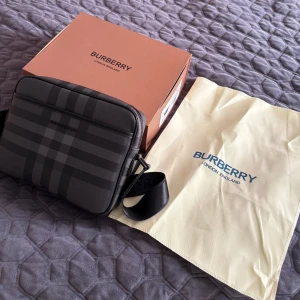 Burberry axelväska - Säljer en snygg Grå/svart axelväska från Burberry. Väskan har svart/grå färgade detaljer, 3styckna dragkedjor fram,mitten,bakre och justerbar axelrem. Perfekt storlek för dem flesta passar jätte bra på sommrarna speicllet utomlands. Endast testad (helt ny) först till kvarn 3500kr