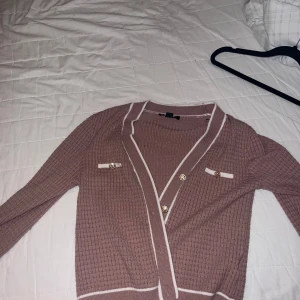 Rosa kofta från Amisu med vita detaljer - Säljer en ribbad rosa kofta från Amisu i storlek XL. (Passar för tjejer med storlek M) Koftan har långa ärmar, vita ränder längs kanterna och dekorativa knappar framtill samt vid fickorna. Perfekt för lager-på-lager och en snygg vardagslook.