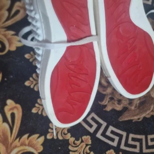 Vita sneakers från Christian Louboutin med röda sulor - Säljer ett par vita sneakers från Christian Louboutin med ikoniska röda sulor och nitar på sidorna. Skorna har snörning och en fransk flagga på insidan av plösen. Perfekta för dig som vill sticka ut med exklusiva detaljer.