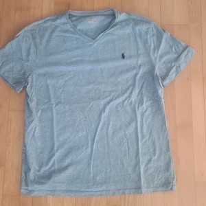 Grå v-ringad t-shirt från Polo Ralph Lauren - Säljer en ljusblå v-ringad t-shirt från Polo Ralph Lauren med den klassiska broderade loggan på bröstet. T-shirten har korta ärmar och är i mjuk bomull, perfekt för en avslappnad stil. Tröjan är I storlek L men sitter minst sagt lika bra på M. Perfekt inför sommaren och bara till ett pris på 299!!! Vilket kap! Orginalpris ligger på 899kr. Priset är inte hugget i sten, kan förhandla