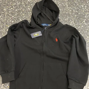 Svart hoodie från Polo Ralph Lauren - Svart hoodie från Polo Ralph Lauren med dragkedja och huva. Klassisk röd logga broderad på bröstet och två fickor framtill. Perfekt för en avslappnad stil och enkel att matcha med det mesta.