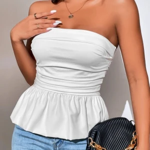 Vit axelbandslös topp med volang - Snygg vit axelbandslös topp med figurnära passform och volang nertill. Perfekt att styla med jeans eller kjol för en trendig look. Enkel och stilren design som passar till många tillfällen.