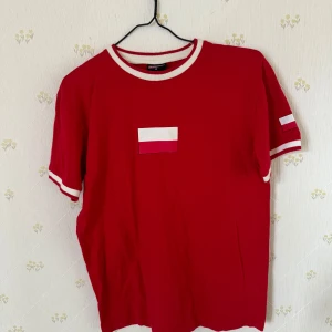 Röd t-shirt från AM Sportia - Röd t-shirt från AM Sportia med vita detaljer vid krage och ärmslut. På bröstet och ena ärmen finns en liten polsk flagga och på ryggen står det 'POLSKA' i vitt. Perfekt för att visa ditt stöd för Polen!