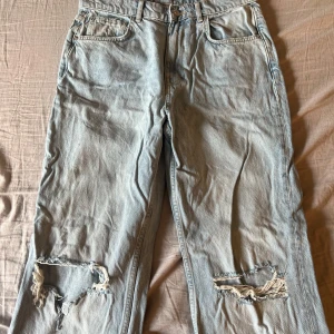 Ljusblå slitna jeans från Gina tricot  - Ljusblå jeans med raka ben och stora slitningar vid knäna från Gina Tricot. Klassisk femficksmodell med normal passform och knappgylf. Perfekta för en avslappnad och trendig look.