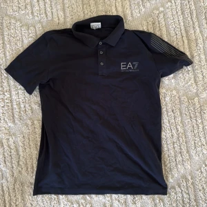 Blå piké tröja från EA7 Emporio Armani - Säljer en blå piké tröja från EA7 Emporio Armani med klassisk krage och tre knappar framtill. T-shirten har korta ärmar och en diskret logga på bröstet. Perfekt för en stilren look.           !!Priset kan diskuteras!!