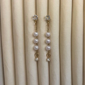 Guldiga örhängen med pärlor och stenar - Säljer ett par eleganta guldiga örhängen med tre vita pärlor på rad och gnistrande stenar upptill och nedtill. Perfekta för att lyfta din look med en touch av lyx och stil. Finns matchande armband till. 