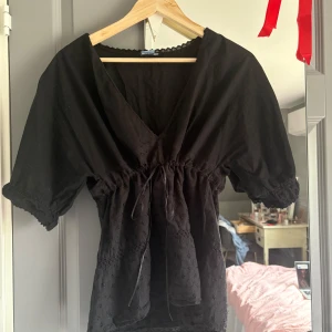 Svart blus med knyt  - Svart blus med v-ringning och knyt i midjan. Blusen har korta puffärmar och spetsdetaljer längs halsringningen. Perfekt för en stilren och feminin look.
